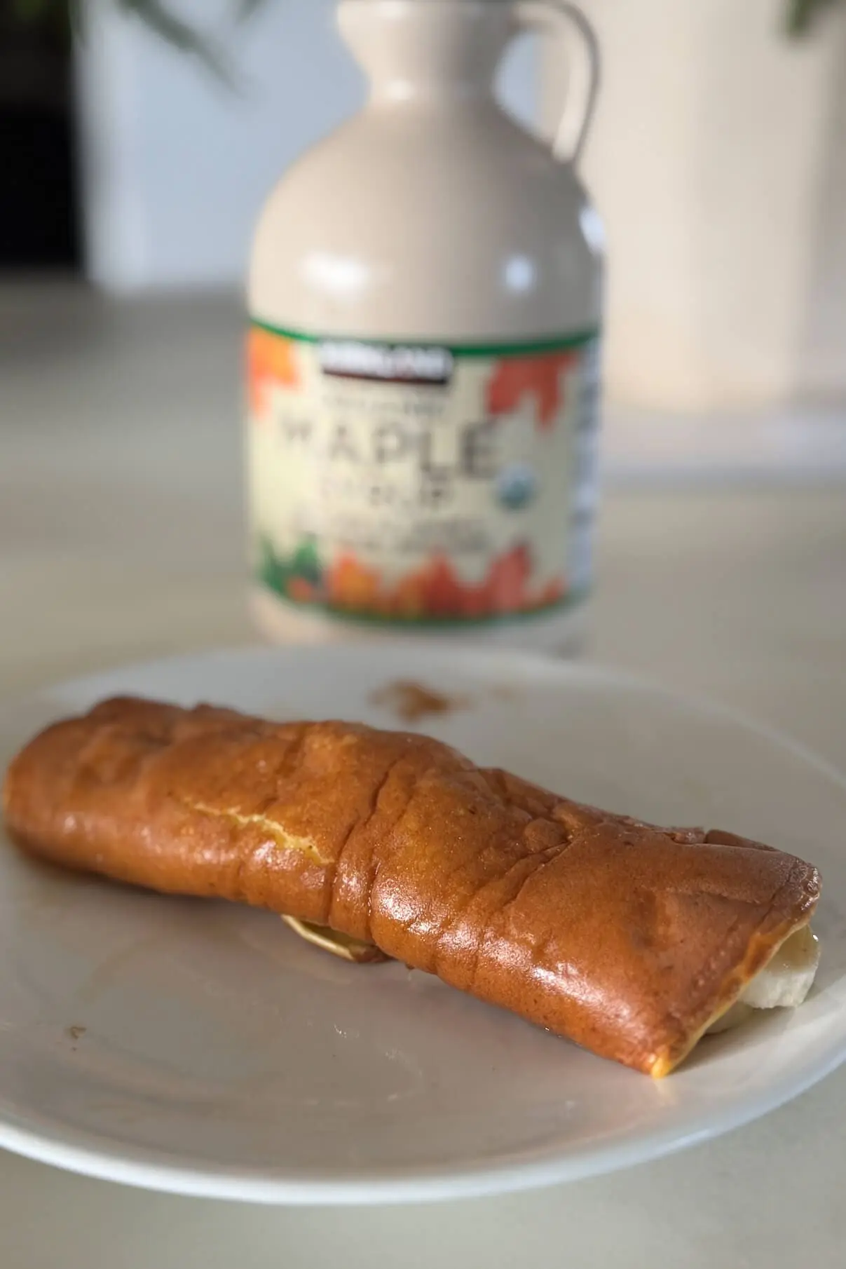 A rolled pancake filled with banana slices, drizzled with syrup, plated on a white dish, with a bottle of organic maple syrup in the background.